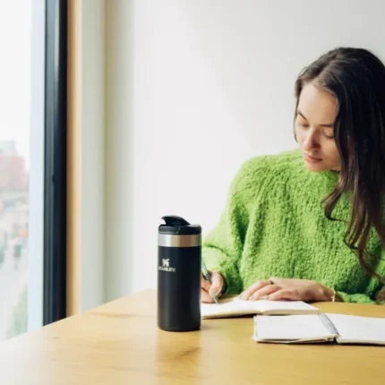 Şişirilmiş termos termosu ile kitap okuyan genç kadın, ev veya ofis ortamında doğal ve konforlu bir çalışma alanında.